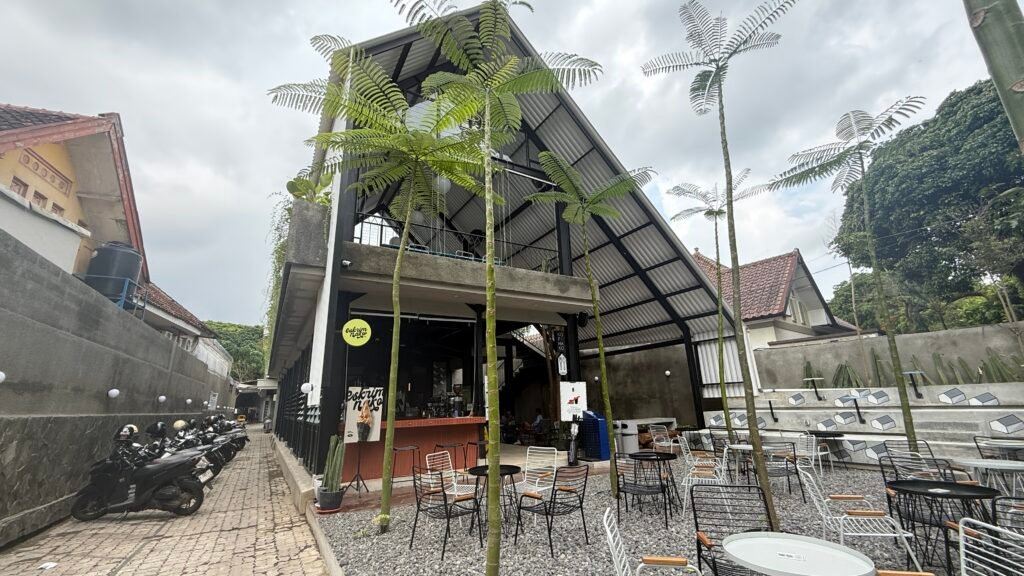 Area outdoor Kopi Nako Malang dengan meja kayu, tanaman hijau, dan cahaya alami.