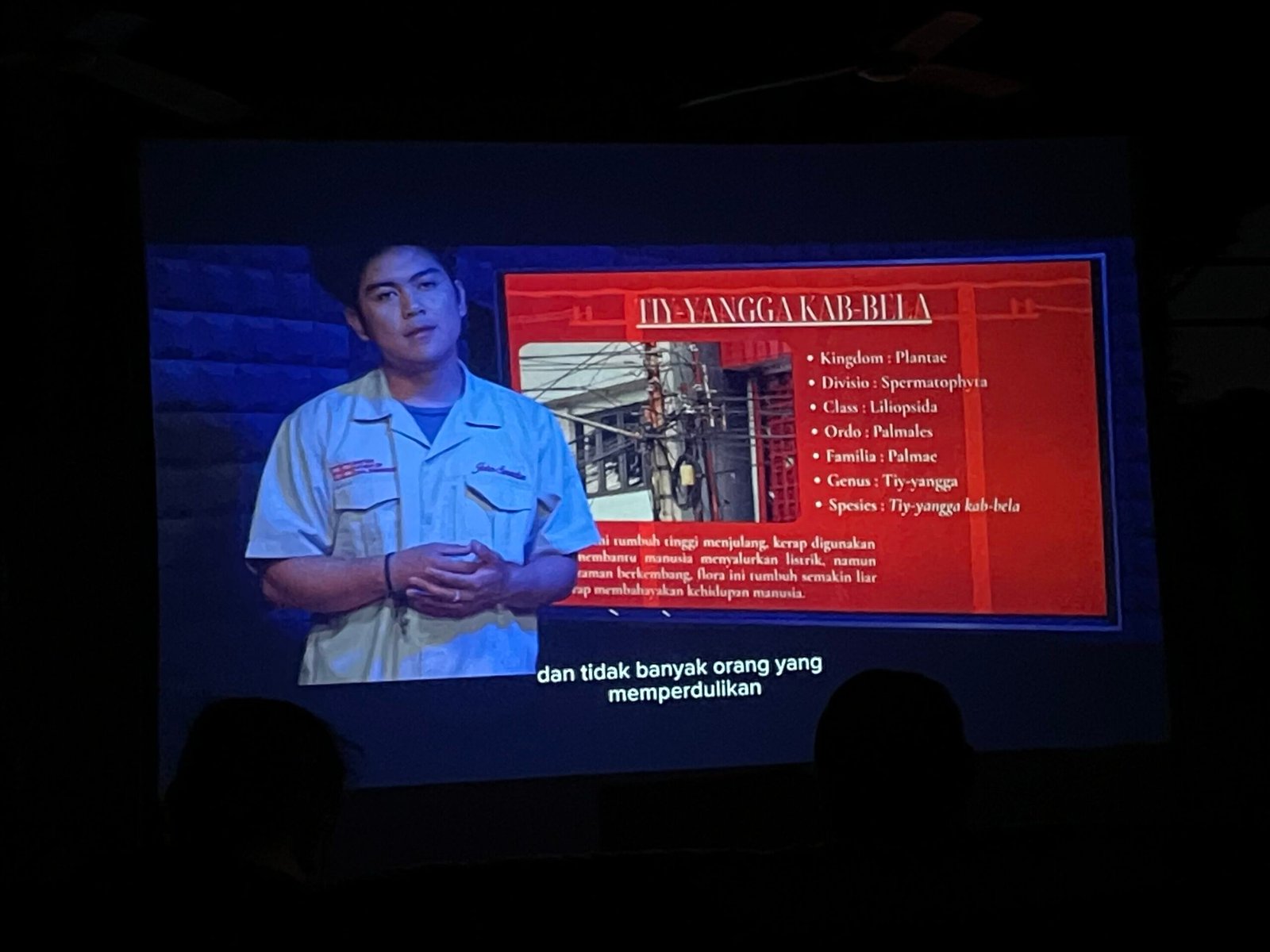 Seorang pemeran dalam film The Presentation tampil di layar proyektor sedang menyampaikan presentasi bertema lingkungan. Di belakangnya terdapat ilustrasi mengenai kabel-kabel semrawut sebagai simbol persoalan lingkungan perkotaan, sementara sejumlah penonton menyaksikan pemutaran film dalam ruangan yang remang-remang.