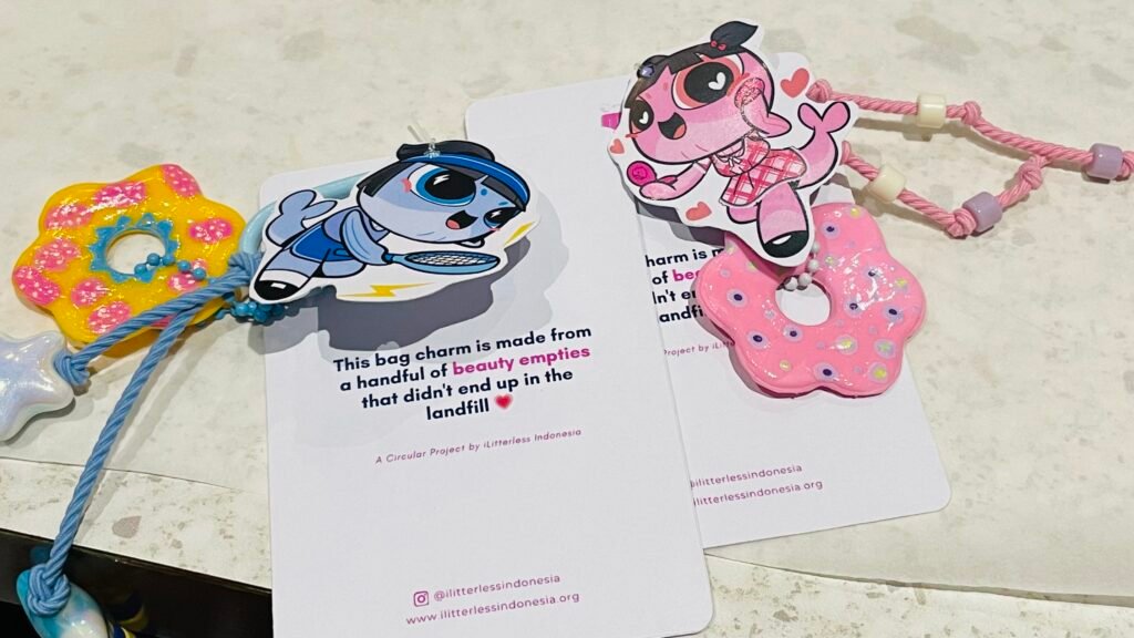 Dua bag charm berwarna biru dan pink hasil daur ulang kemasan produk kecantikan dipajang di atas kartu informasi iLitterless Indonesia. Aksesori tersebut dibuat dari plastik HDPE bekas yang diolah kembali menjadi produk bernilai guna dan estetis.